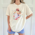 Person wearing a cream t-shirt with a colorful scooter graphic of a girl and cat surrounded by flowers and the text "Be Brave Not Perfect".