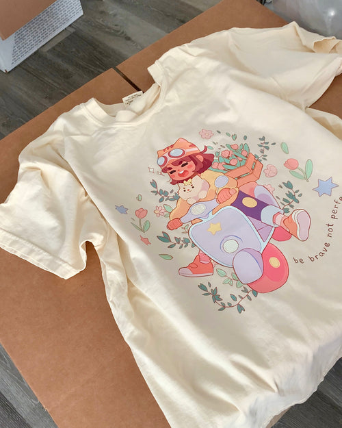Cream-colored “Be Brave Not Perfect” t-shirt featuring scooter girl graphic shown in a flat lay on a cardboard surface.