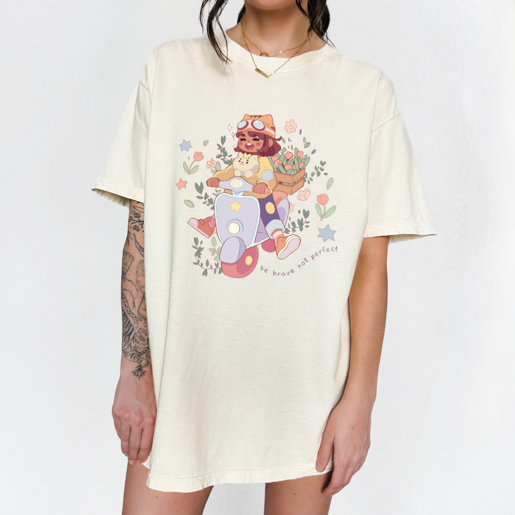 Cream-colored oversized t-shirt with “Be Brave Not Perfect” scooter girl graphic, modeled standing against a white background.