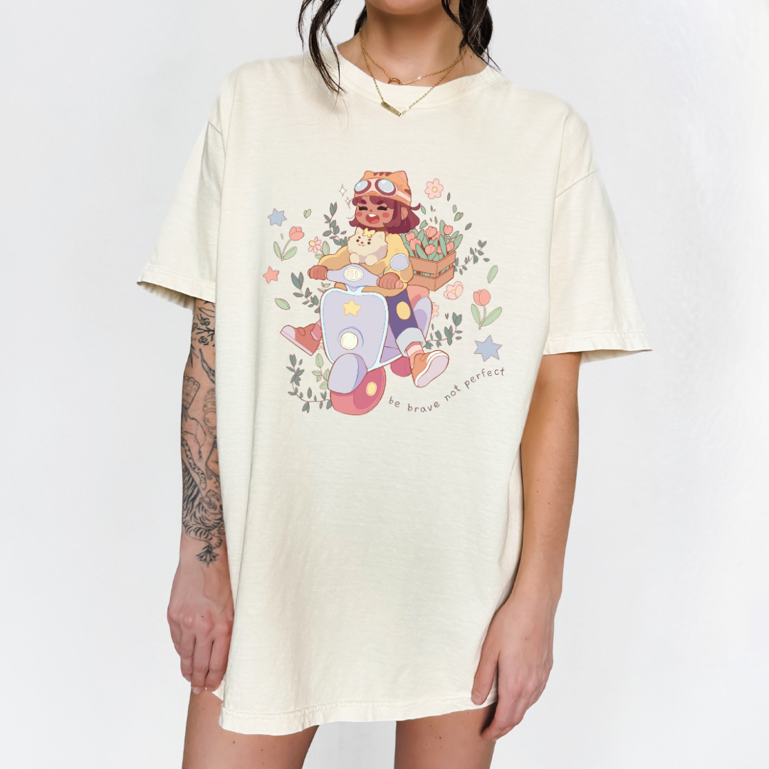 Cream-colored oversized t-shirt with “Be Brave Not Perfect” scooter girl graphic, modeled standing against a white background.