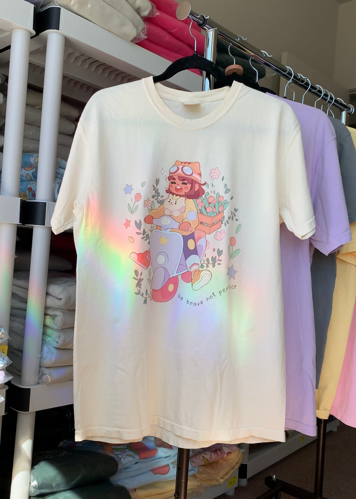 Cream-colored “Be Brave Not Perfect” graphic t-shirt hanging on a clothing rack with rainbow light reflections in a retail display setting.
