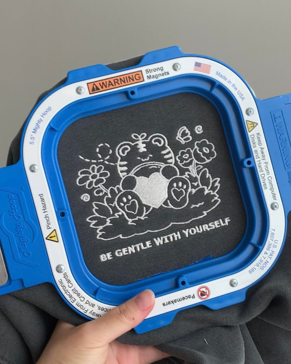 Close-up of in-house embroidery on a charcoal gray sweatshirt showing a gentle character with florals and the phrase “Be Gentle With Yourself,” stitched in white thread.