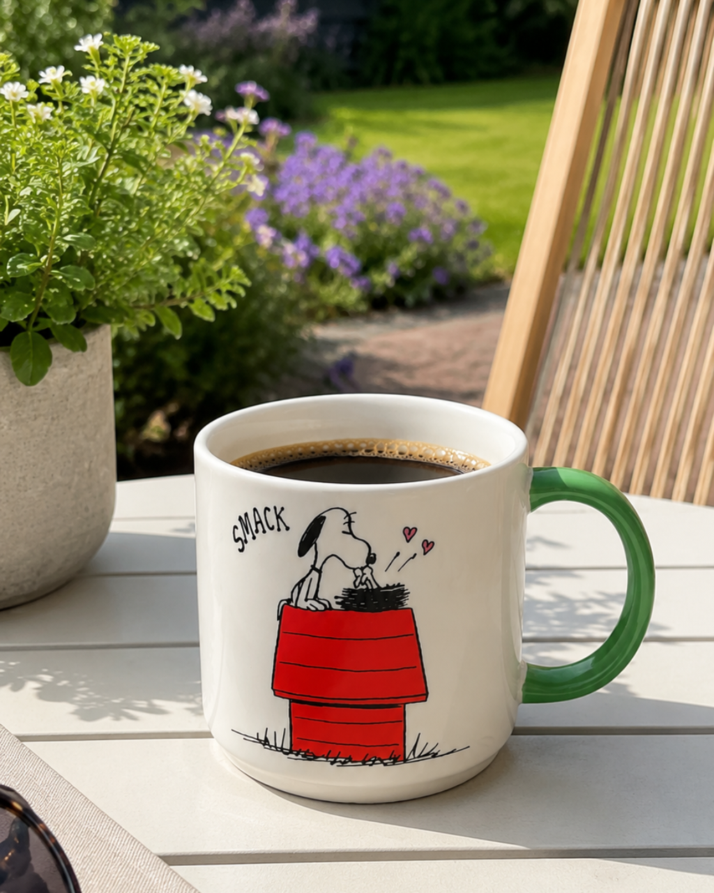 A green-handled Snoopy mug featuring Snoopy kissing Woodstock atop his red doghouse sits on an outdoor garden table surrounded by purple flowers.