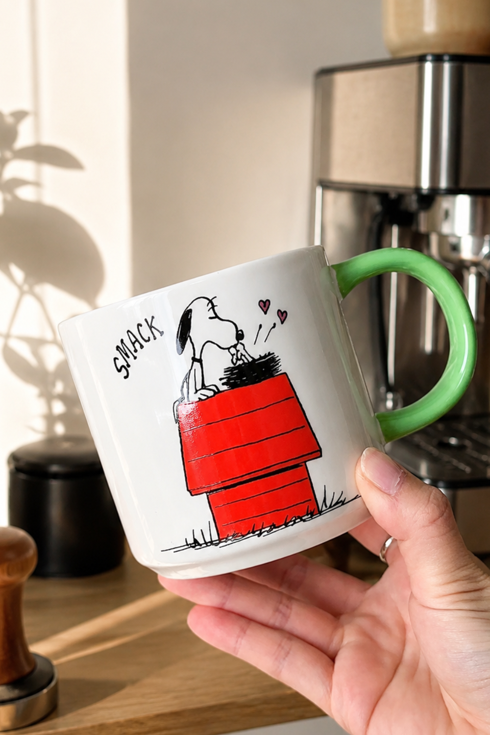 Snoopy and Woodstock Be Kind to All Kinds ceramic mug with green handle displayed in a cozy coffee station with warm morning sunlight.