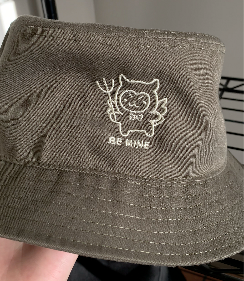 Close-up of an olive green bucket hat held in hand, showing detailed embroidery of a devil-cat character with a pitchfork and "Be Mine" text.
