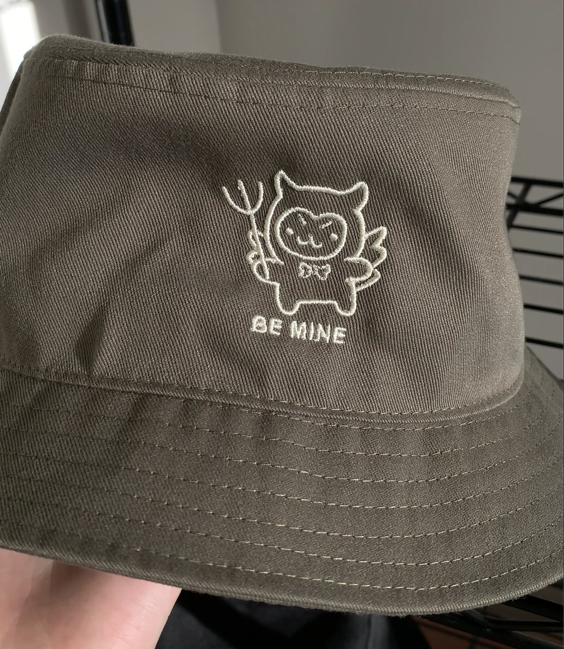 Close-up of an olive green bucket hat held in hand, showing detailed embroidery of a devil-cat character with a pitchfork and "Be Mine" text.
