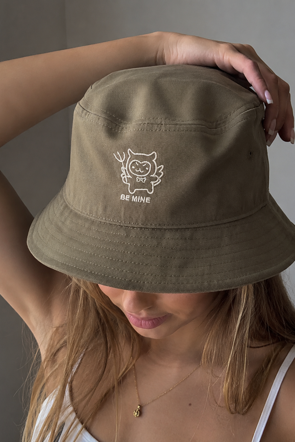 Woman wearing an olive green bucket hat with embroidered "Be Mine" design, holding the brim, close-up shot with a gold necklace visible.
