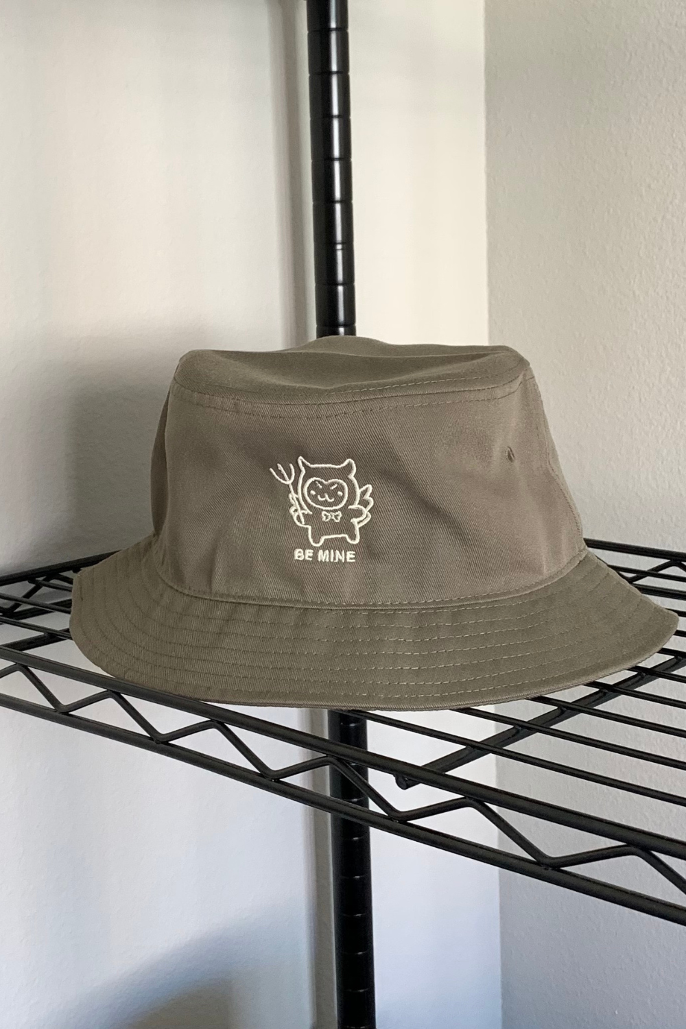 Olive green bucket hat with a small embroidered devil-cat character and "Be Mine" text, displayed on a wire shelf against a white wall.
