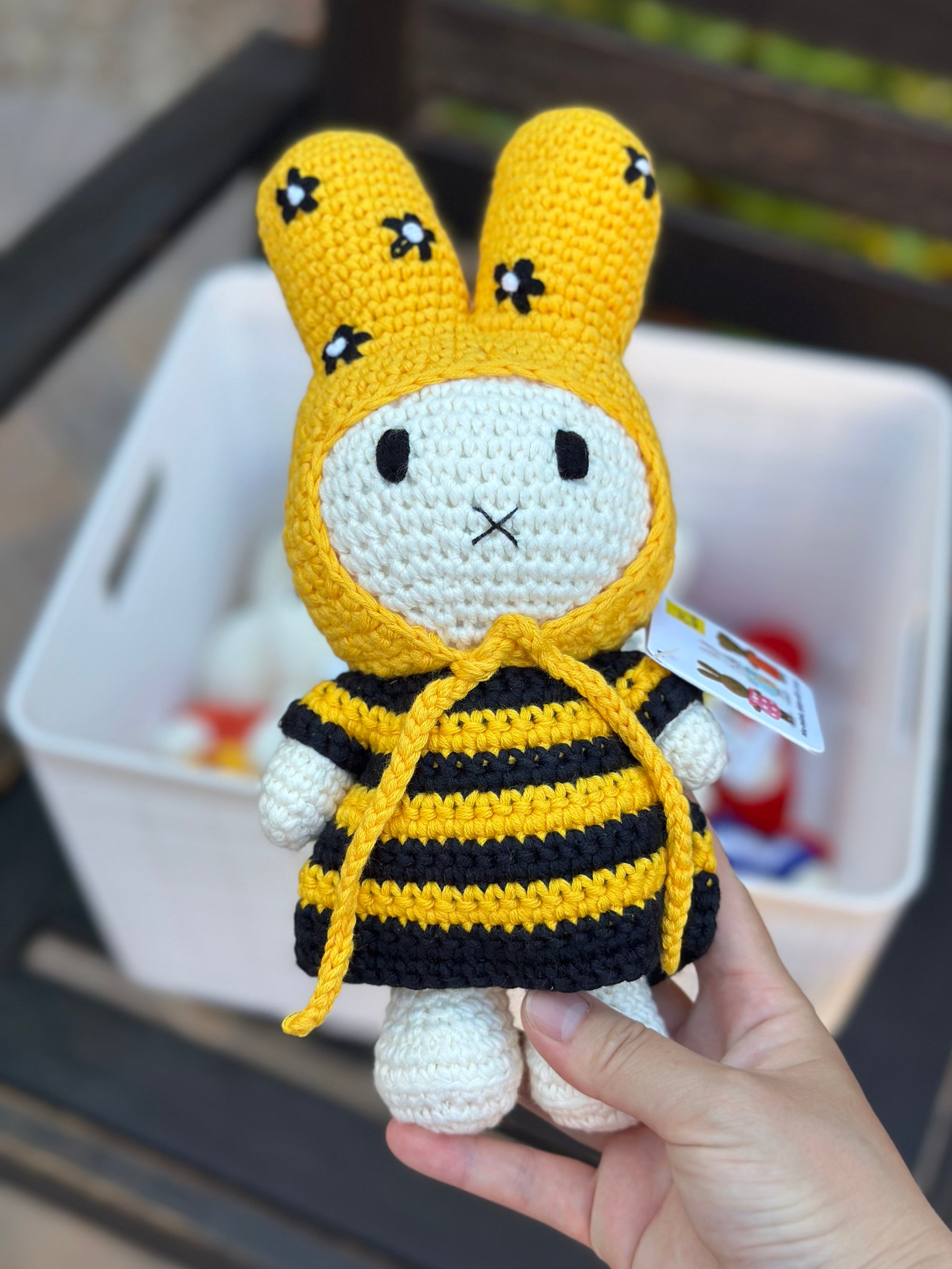 Handmade crochet Miffy plush in a bee-striped dress and yellow bonnet with flowers, held in front of a basket of other crochet dolls.