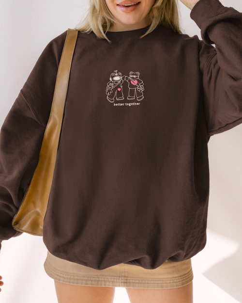 Model wearing a brown embroidered sweatshirt with two characters and the phrase “Better Together,” styled for everyday casual wear.