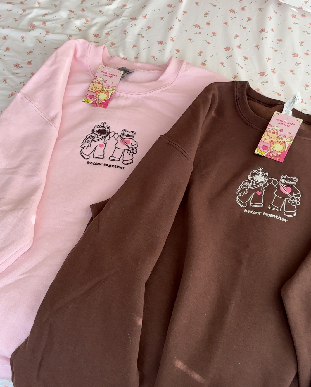 Pink and brown embroidered crewneck sweatshirts with two hand-holding characters and the words “better together,” laid flat on a floral bedspread.