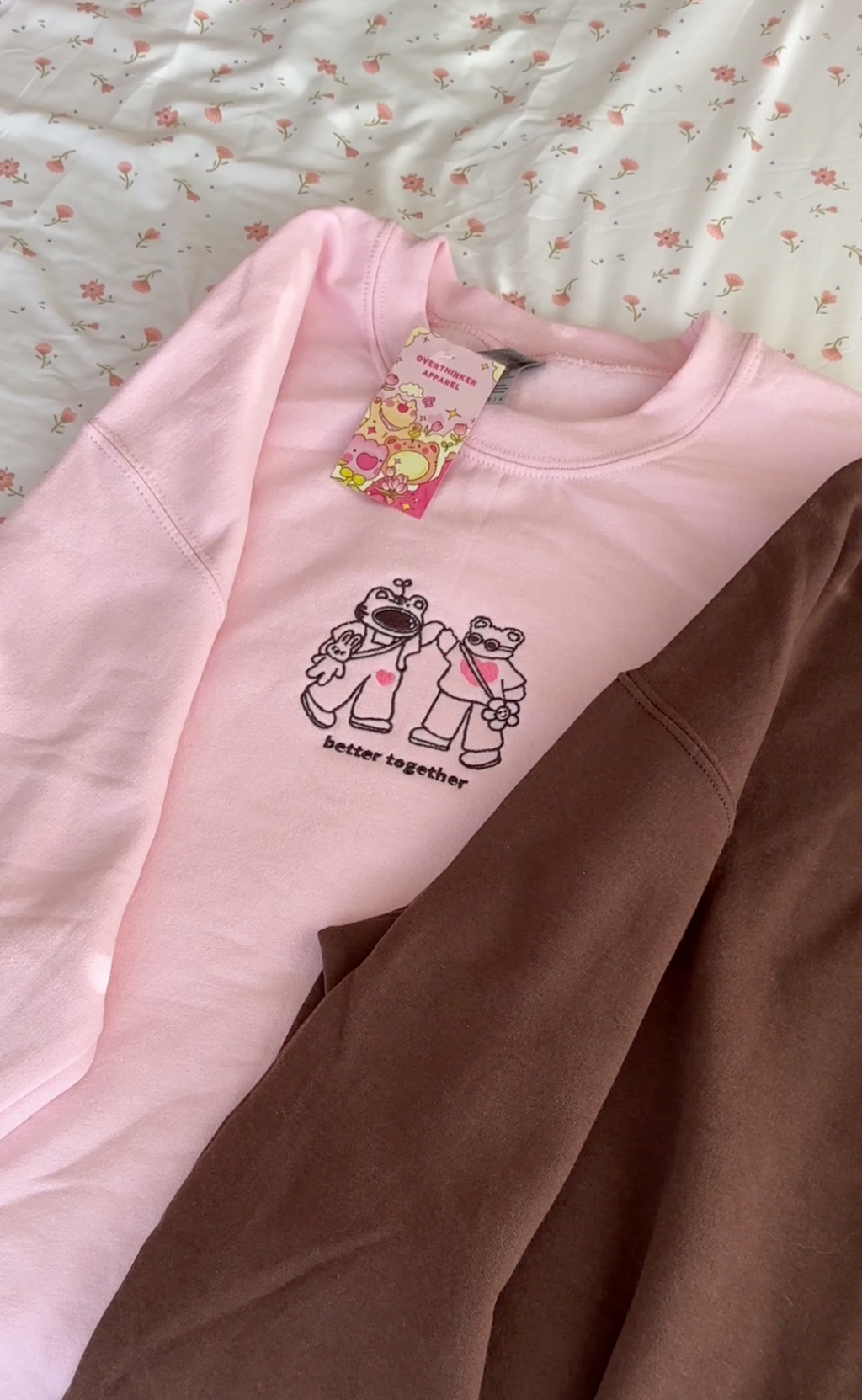 Light pink embroidered sweatshirt with a minimalist character design and “better together” text, shown laid flat on a bed.