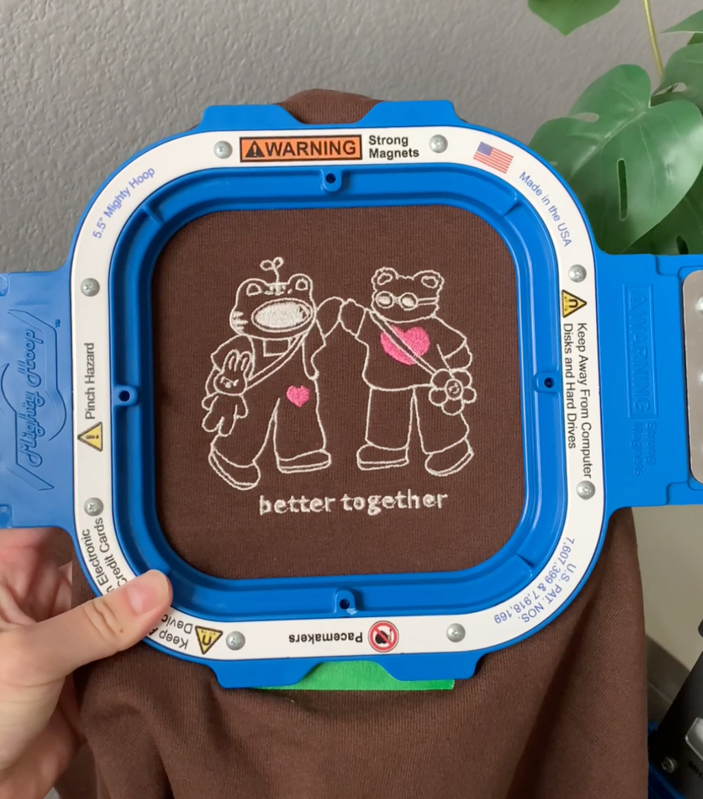 Close-up of embroidered “better together” design on a brown sweatshirt, showing two stitched characters holding hands with heart accents.