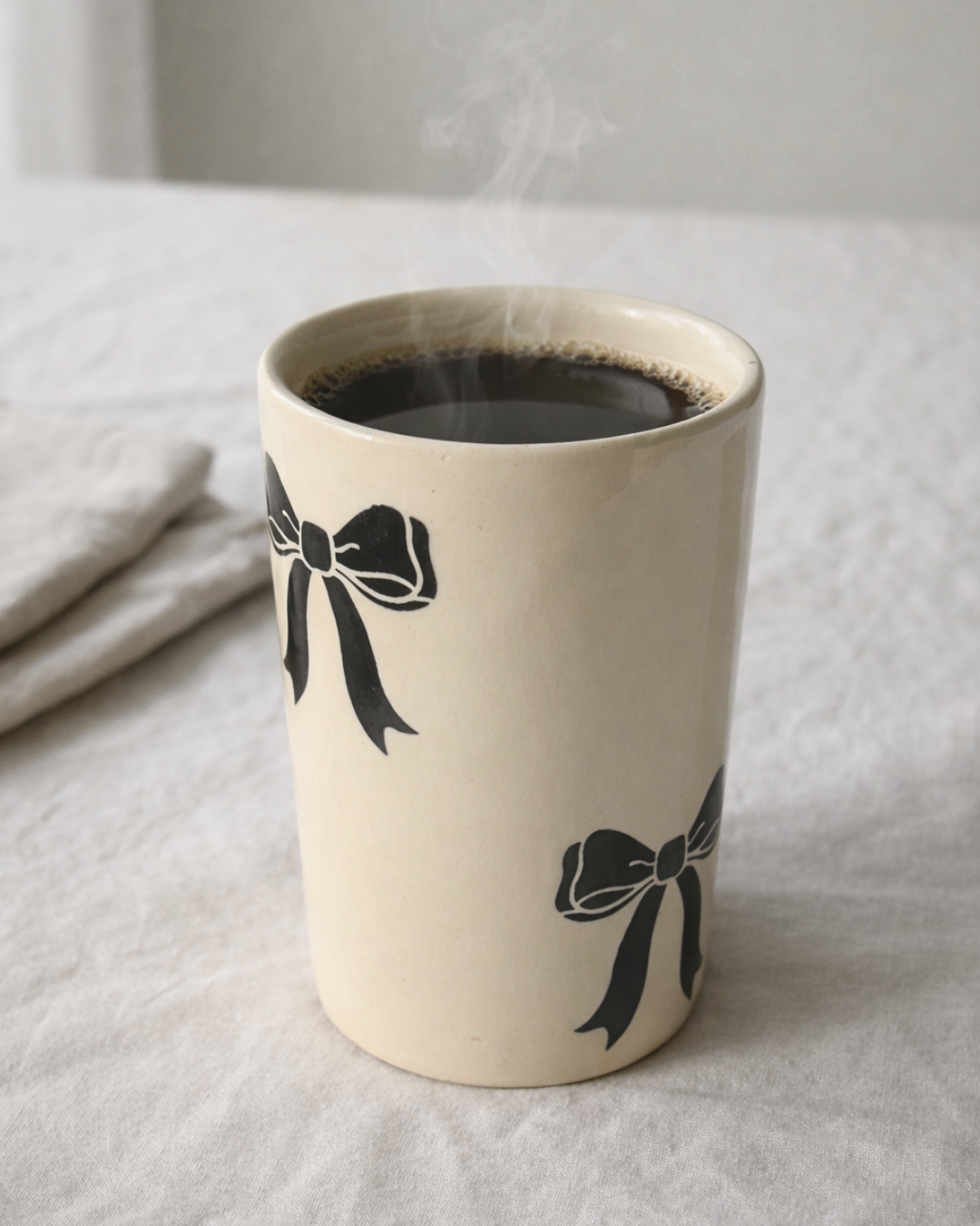Handmade ceramic bow coffee mug with black bow design filled with hot coffee on linen cloth, aesthetic ceramic mug for cozy morning coffee ritual