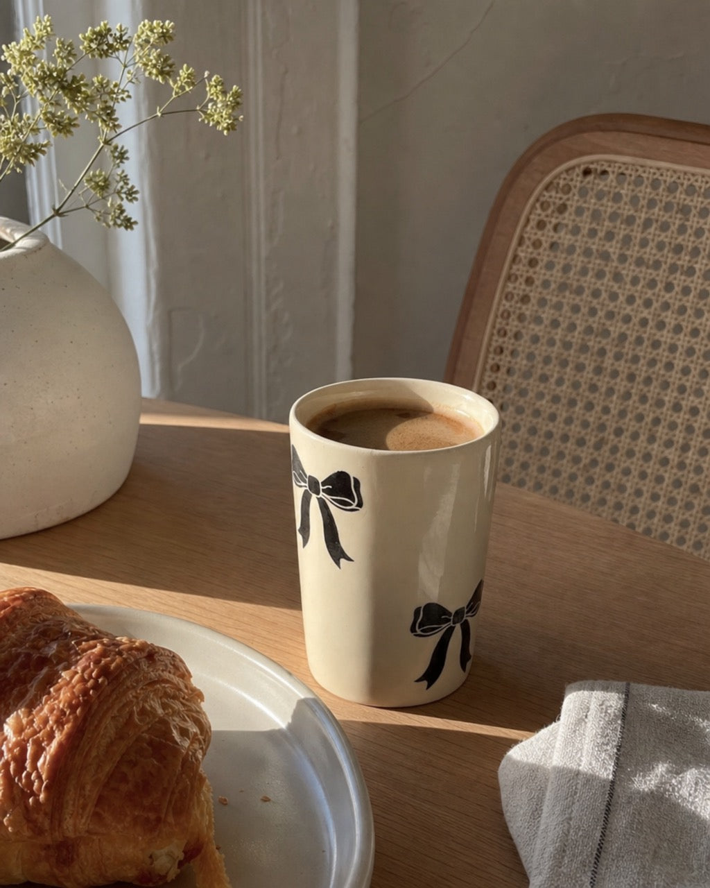 A beige ceramic coffee mug with black bow illustrations sits on a wooden table in a cozy cafe-like setting, next to a croissant on a plate and a white vase with dried flowers.