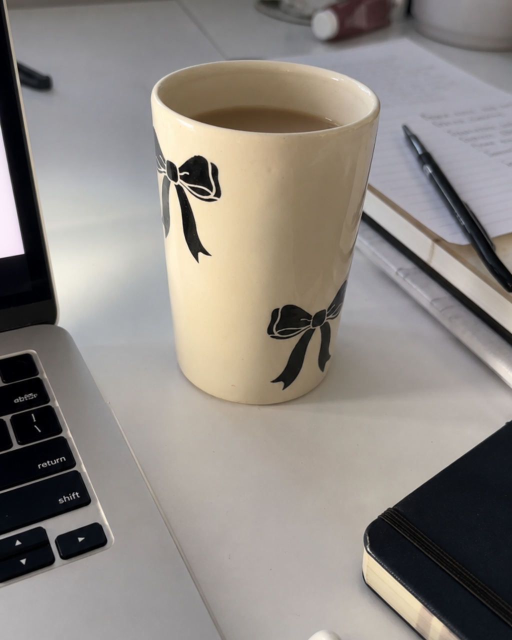 A black bow ceramic mug filled with coffee on a white desk next to a laptop, notebook, and pen.