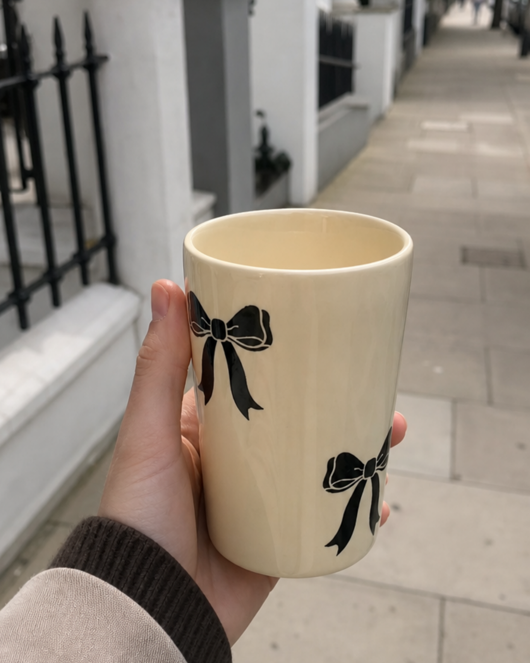 A hand-painted black bow ceramic mug held up on a city sidewalk, black bow design visible against cream stoneware.