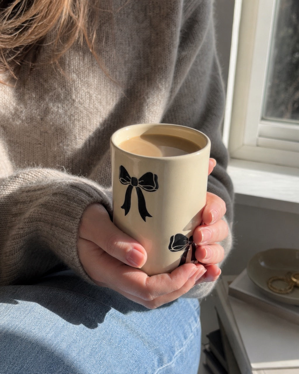 A person in a cozy gray sweater holds a beige ceramic mug with black bow illustrations, filled with a light brown beverage, near a bright window.