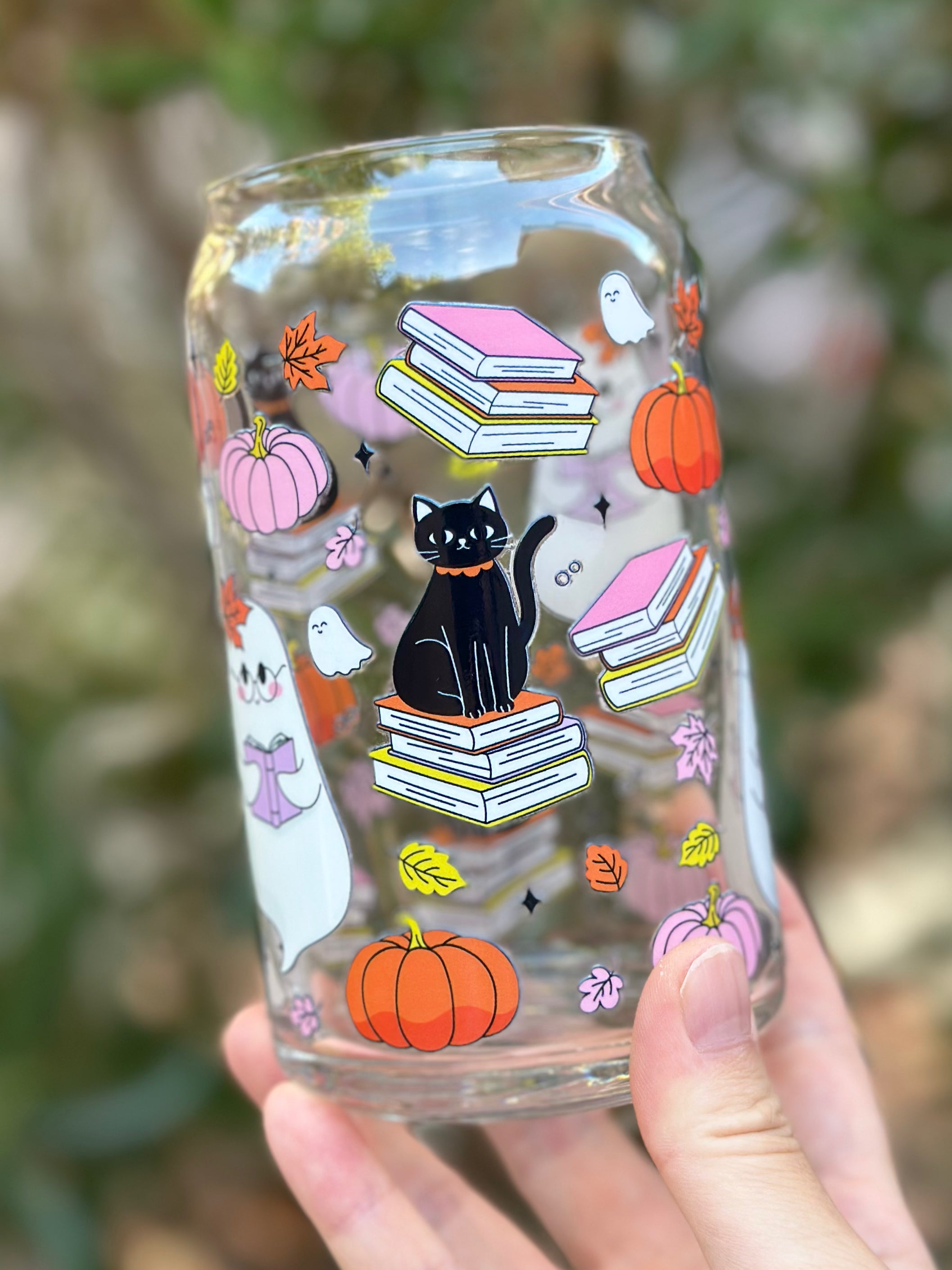 Clear glass can cup decorated with a black cat sitting on books, a ghost reading, pumpkins, pink books, and autumn leaves.