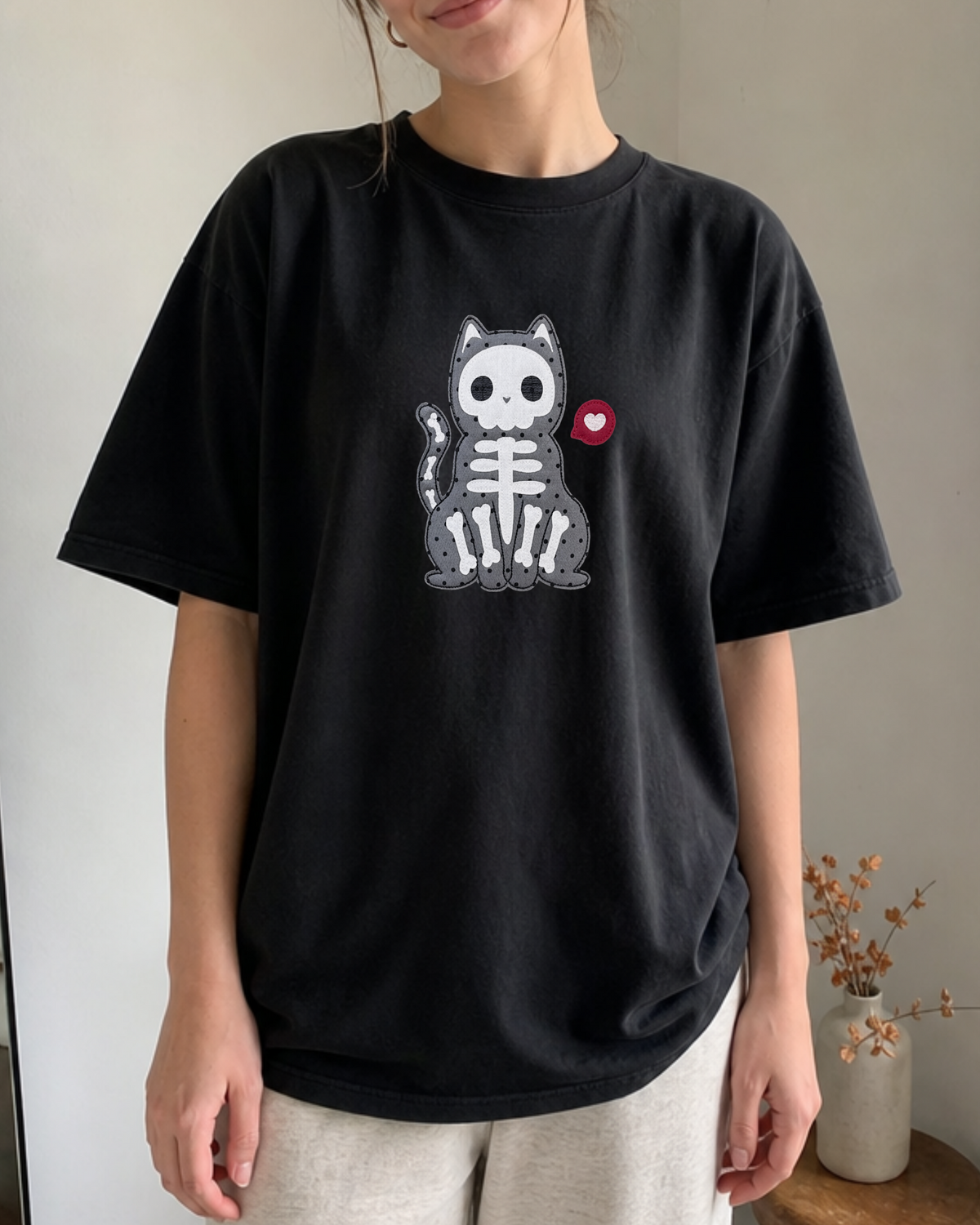 A person wearing a black short-sleeve graphic T-shirt with a gray cat skeleton design and a small red heart on the chest, paired with light beige pants in an indoor setting.