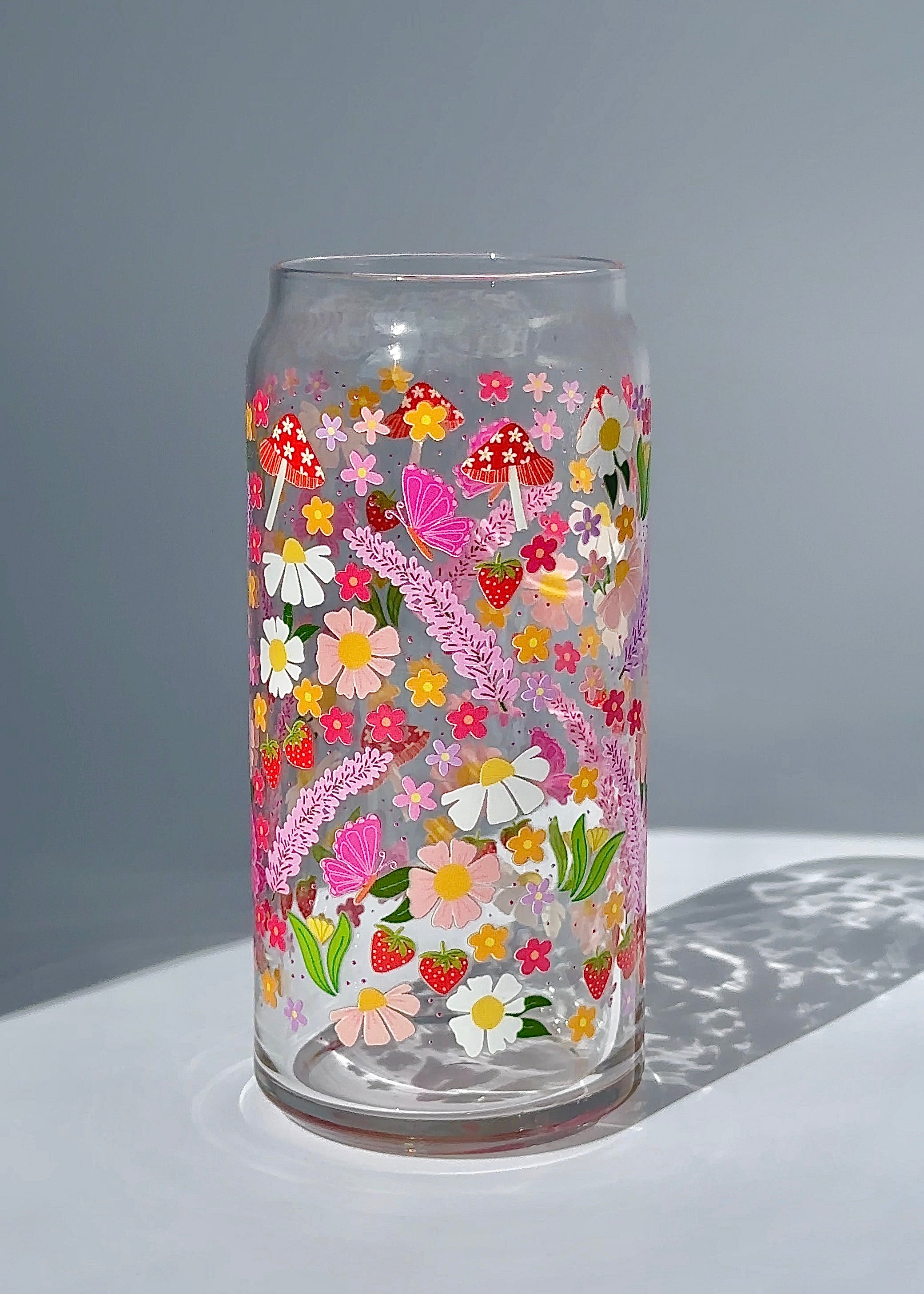 Clear 20 oz glass can featuring a blooming garden illustration with colorful flowers, strawberries, mushrooms, and butterflies, styled in bright natural light.