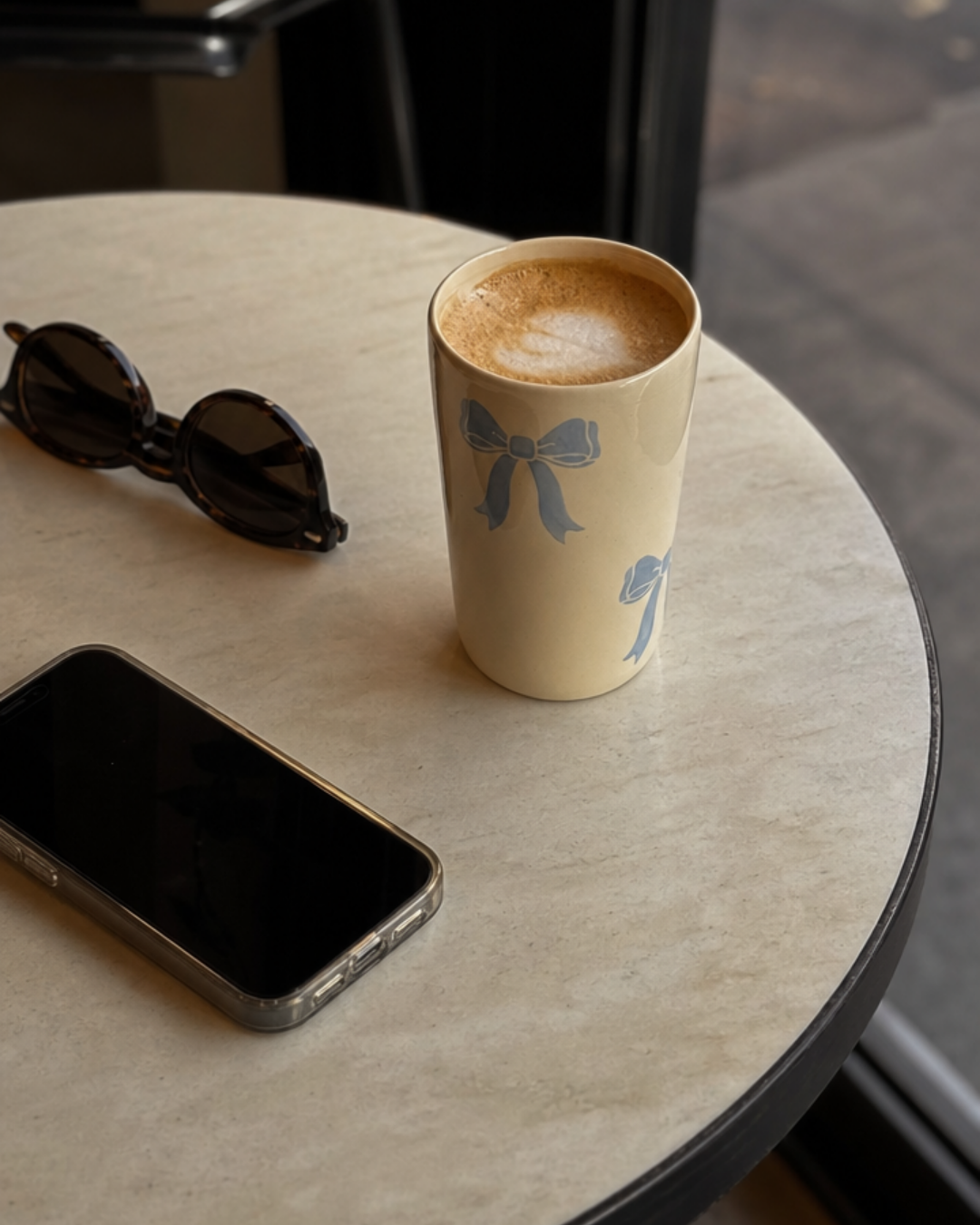 A beige ceramic coffee mug with a blue bow print sits on a round stone table next to sunglasses and a smartphone.