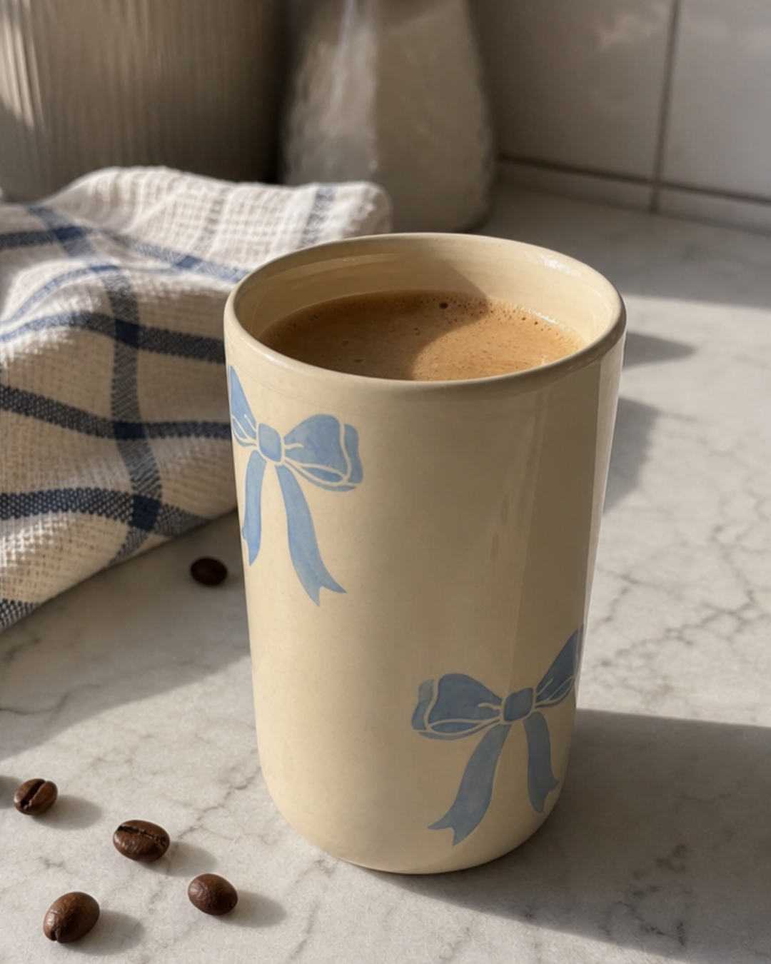 A cream-colored ceramic mug with light blue bow patterns, filled with coffee, set on a marble countertop with coffee beans and a blue-and-white checkered cloth nearby.