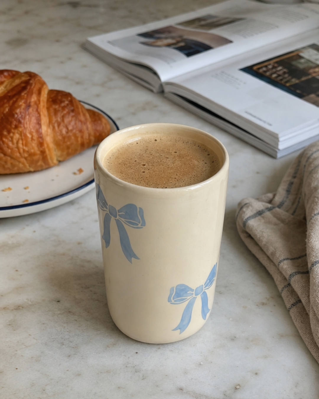 A cream-colored ceramic mug with light blue bow patterns, filled with coffee, set on a marble countertop next to a croissant and an open magazine.