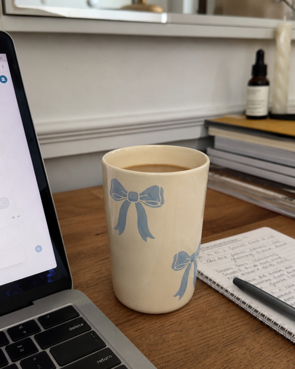 A cream-colored ceramic mug with a light blue bow pattern sits on a wooden desk next to an open laptop, a notebook, and a pen, with a blurred home office background.
