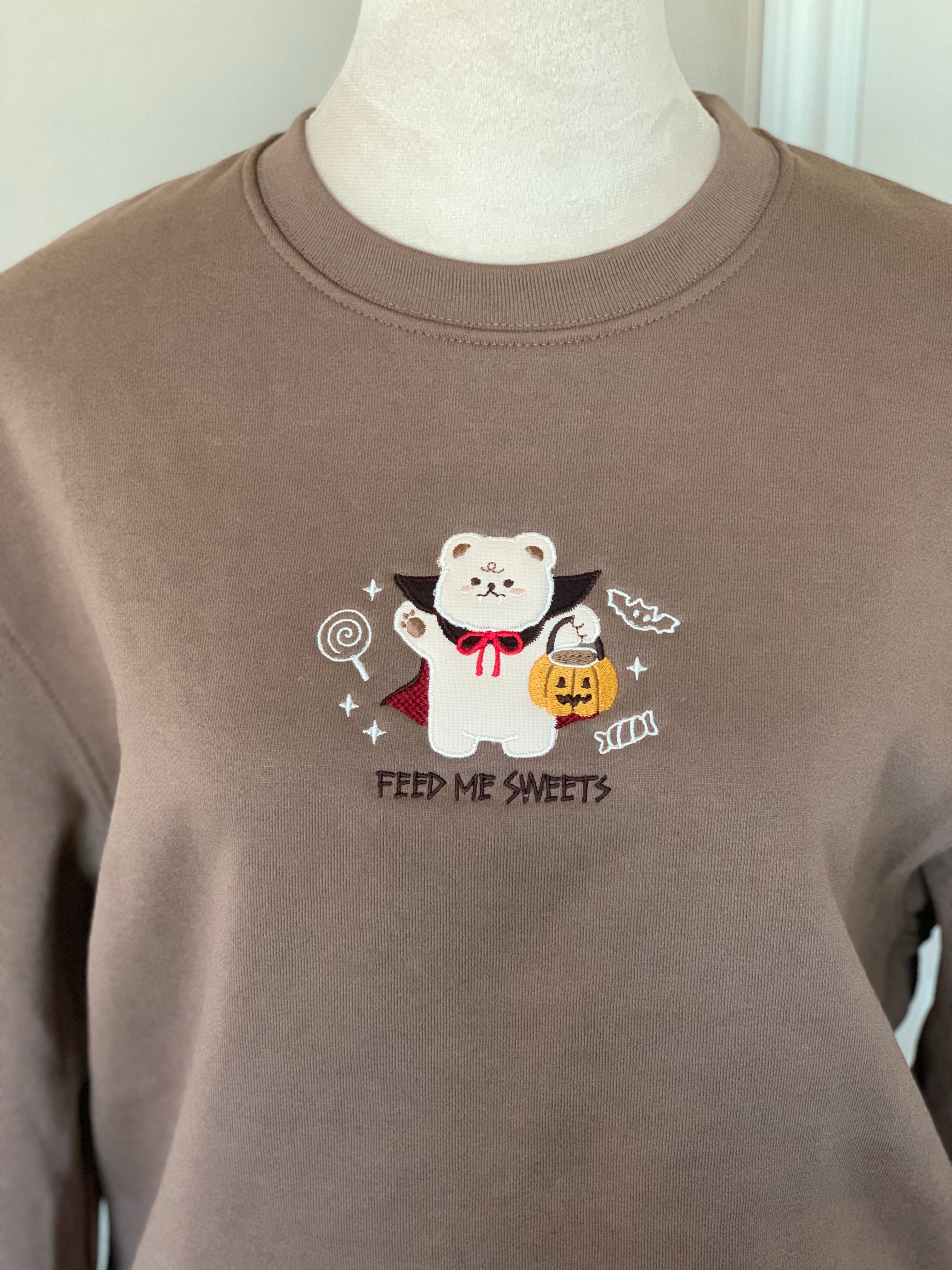 Front close-up of brown sweatshirt with embroidered vampire bear in red cape holding pumpkin bucket, surrounded by candy.