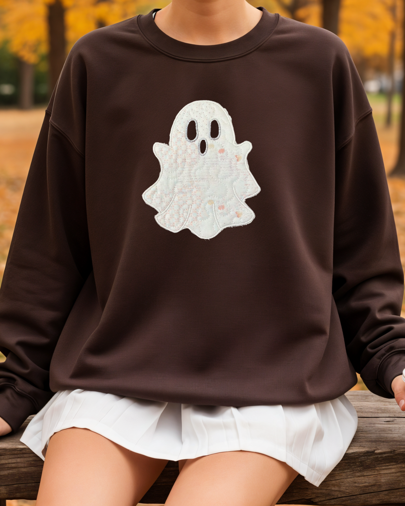 Person sitting outdoors in autumn wearing a brown sweatshirt with large white embroidered ghost design.