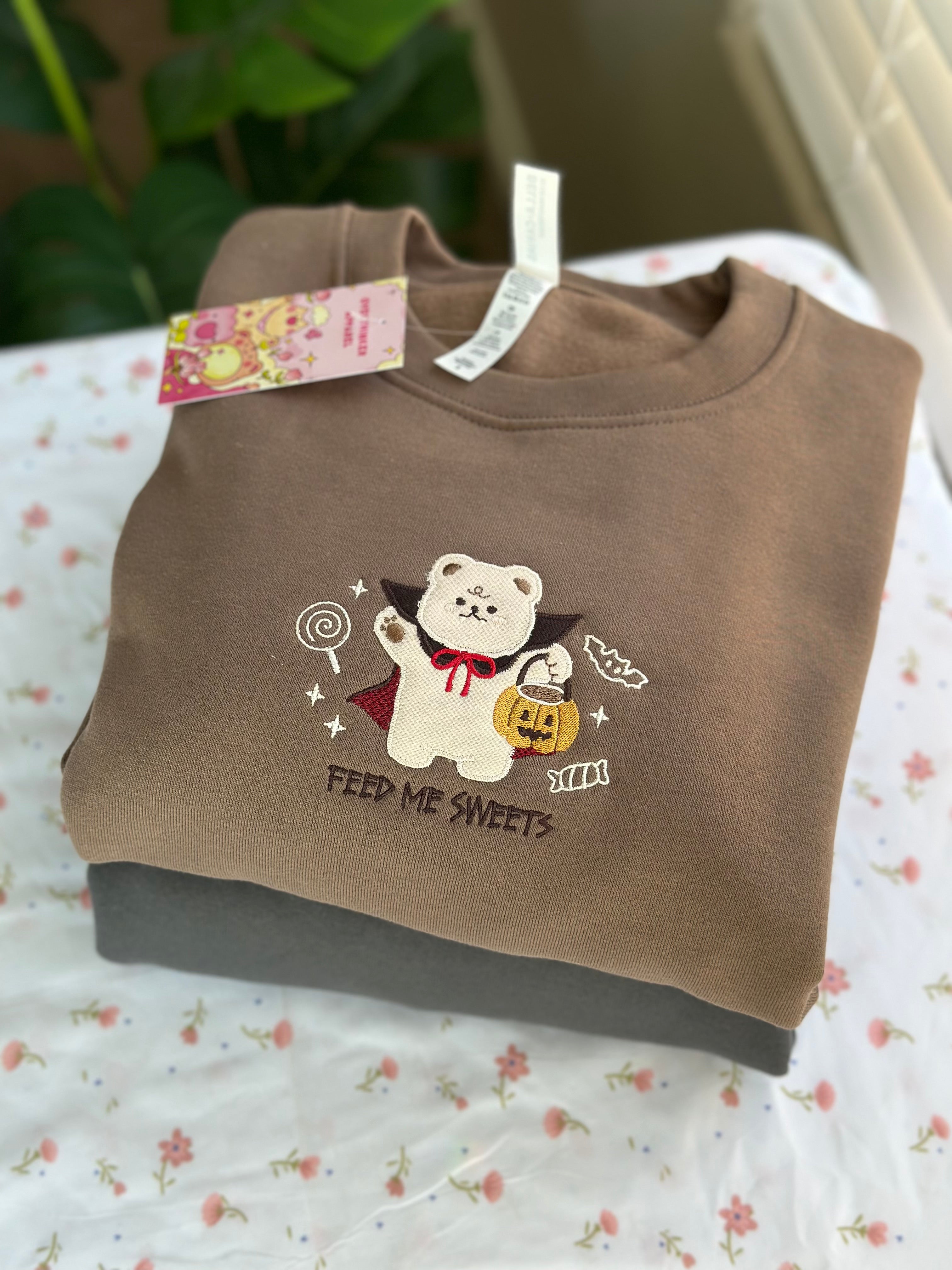 Folded brown sweatshirt with embroidered vampire bear holding a pumpkin bucket and candy, text reads “Feed Me Sweets.”