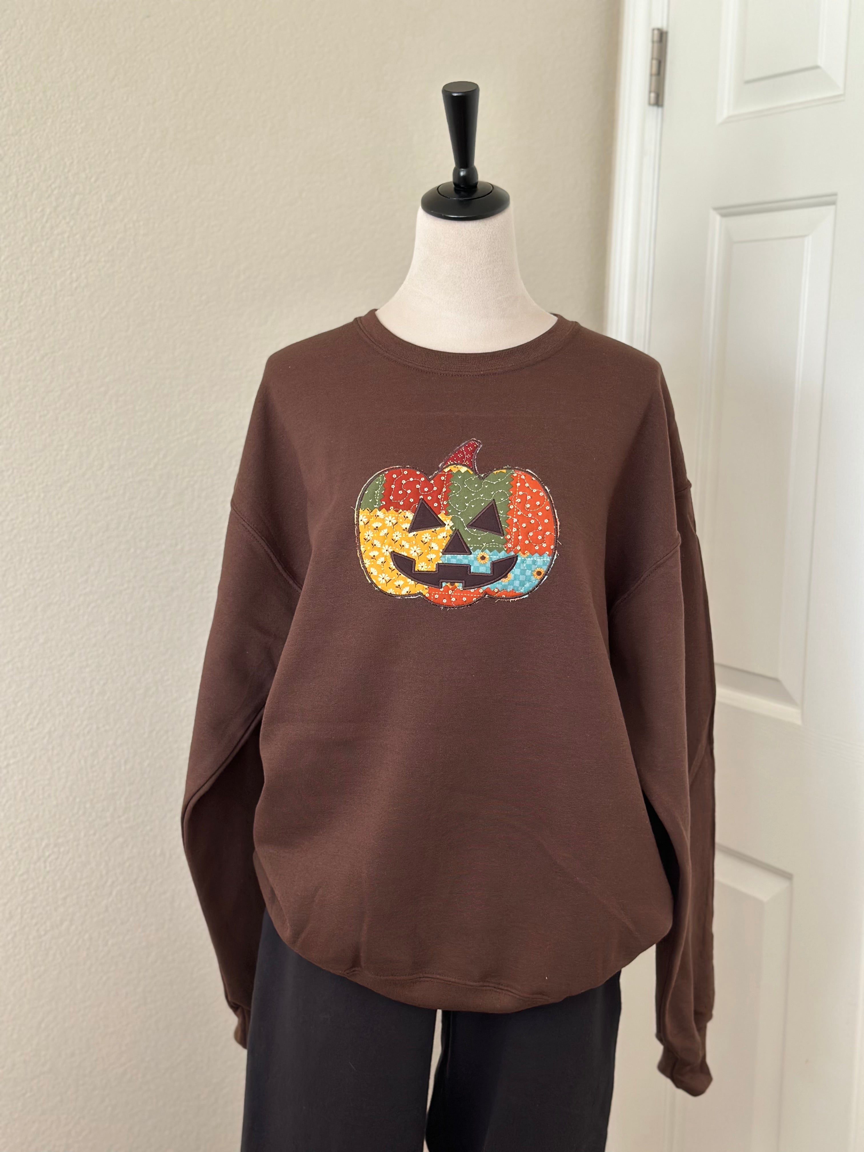 Brown sweatshirt displayed on mannequin with large colorful patchwork jack-o’-lantern applique on the front.