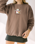 Person wearing a brown sweatshirt with embroidered vampire bear holding pumpkin candy bucket and “Feed Me Sweets” text, styled with a skirt and gold jewelry.