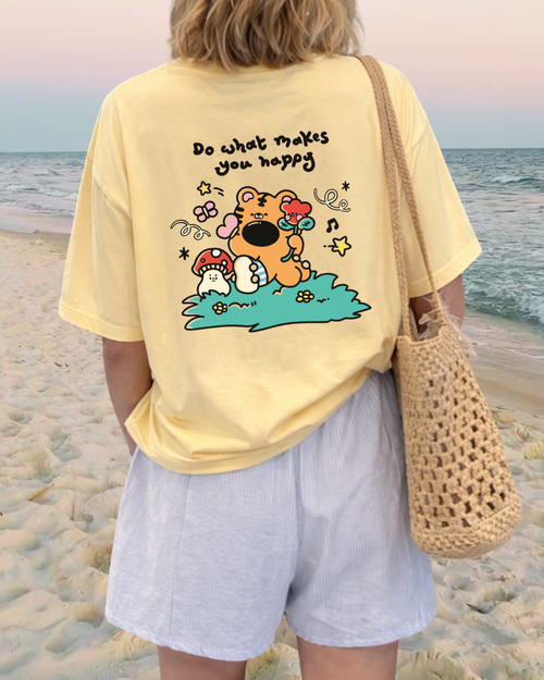 Woman wearing a butter yellow graphic tee with a cute bear illustration and “Do What Makes You Happy” quote printed on the back, styled with relaxed shorts at the beach.