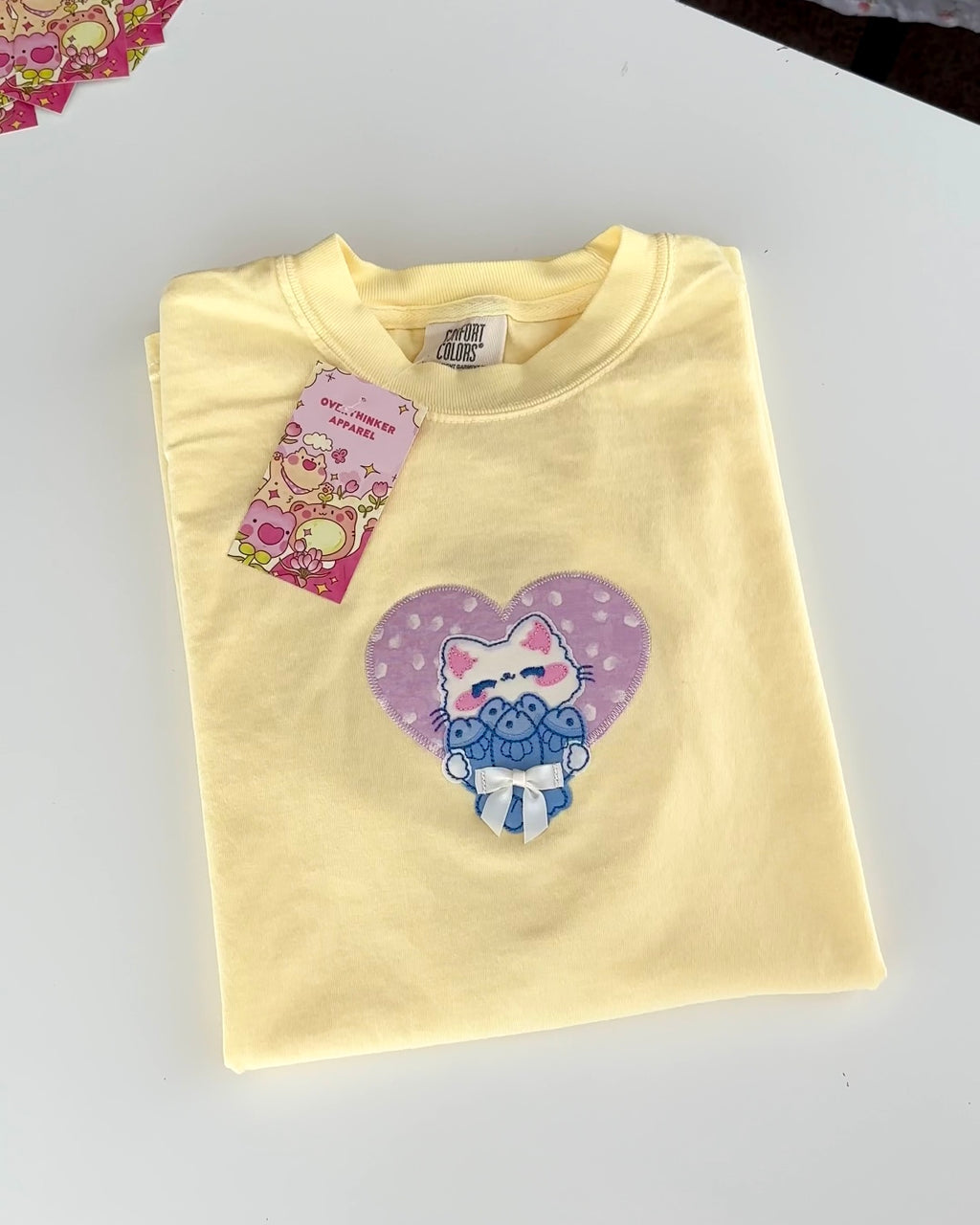 Flat lay of pastel yellow embroidered cat patchwork t-shirt featuring a lilac heart and satin bow detail.