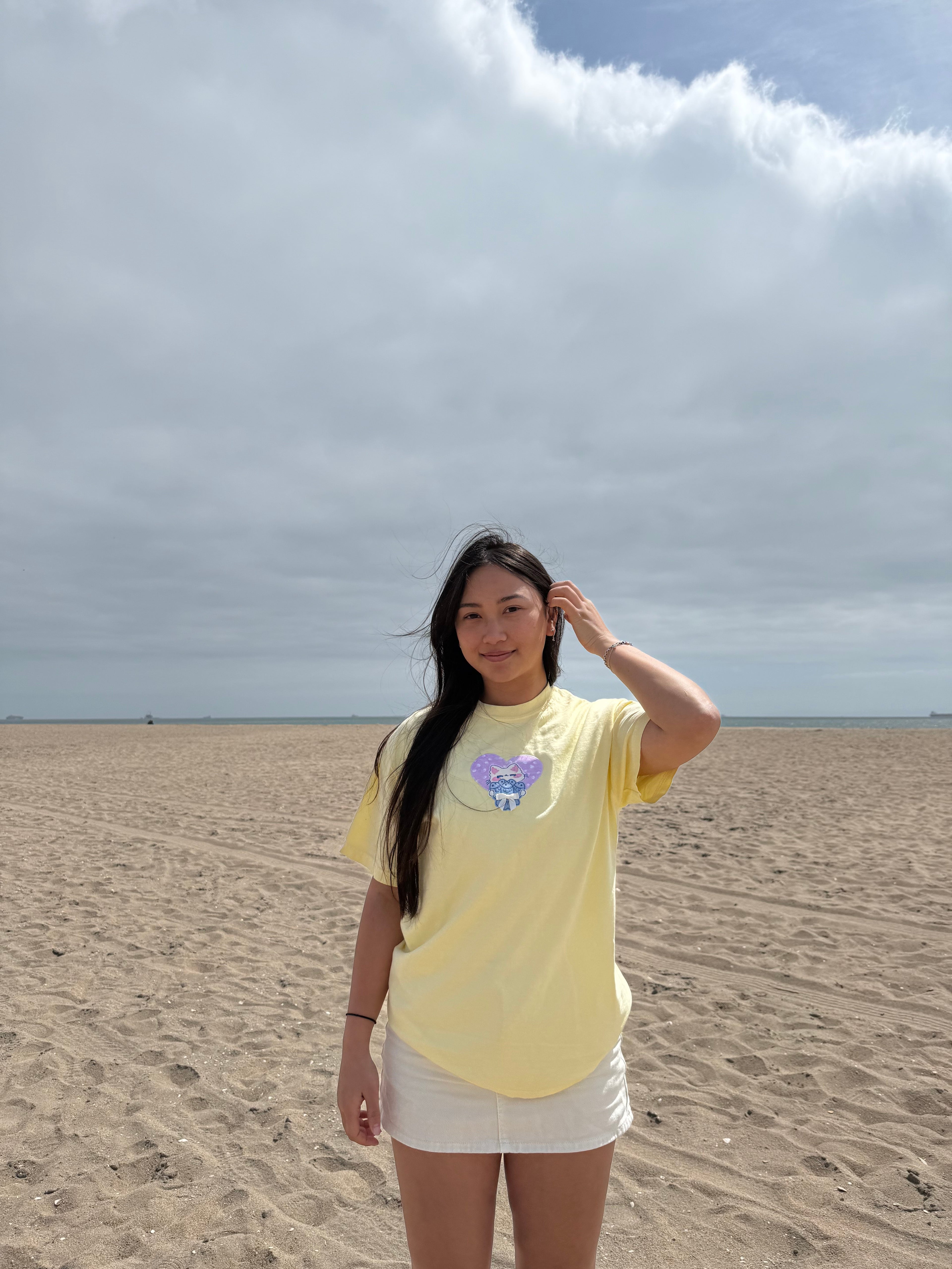Model wearing a pastel yellow embroidered cat patchwork t-shirt at the beach, featuring a white cat holding fish inside a lilac heart.