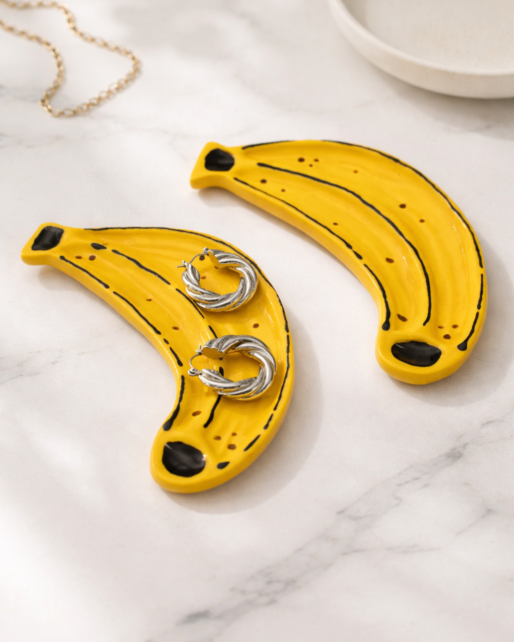 Ceramic banana jewelry dish with gold earrings on marble surface, handmade fruit shaped ring holder for vanity organization and quirky home decor