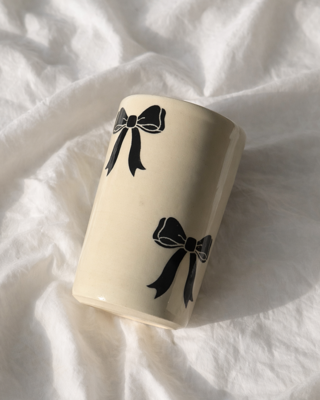 Handmade ceramic bow mug with black bow design resting on soft linen fabric, aesthetic ceramic coffee cup for cozy coffee rituals and feminine kitchen decor