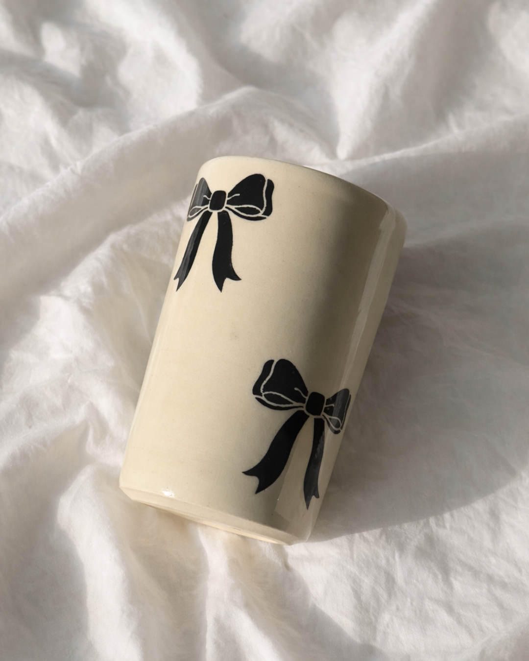Handmade ceramic bow mug with black bow design resting on soft linen fabric, aesthetic ceramic coffee cup for cozy coffee rituals and feminine kitchen decor