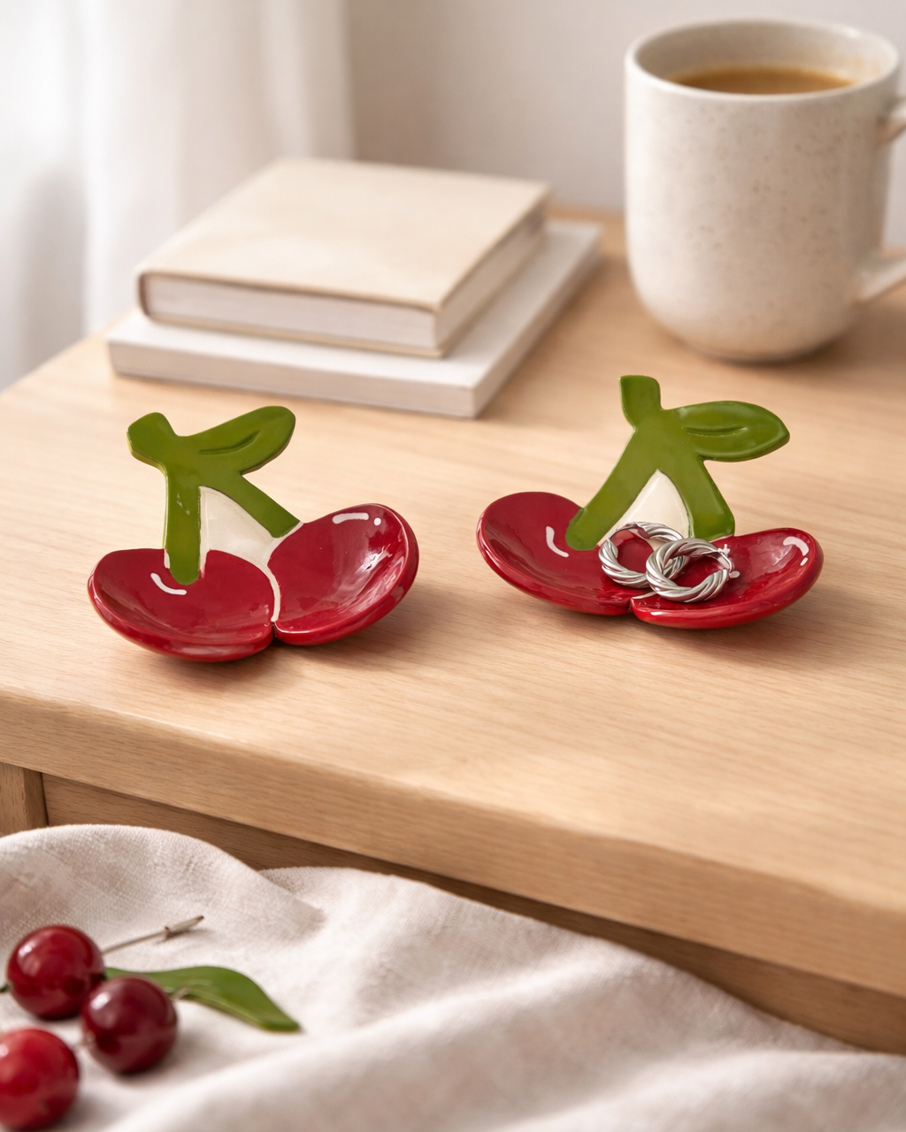 Handmade ceramic cherry ring dish holding silver earrings on bedside table next to coffee mug and books, fruit shaped jewelry holder for nightstand decor