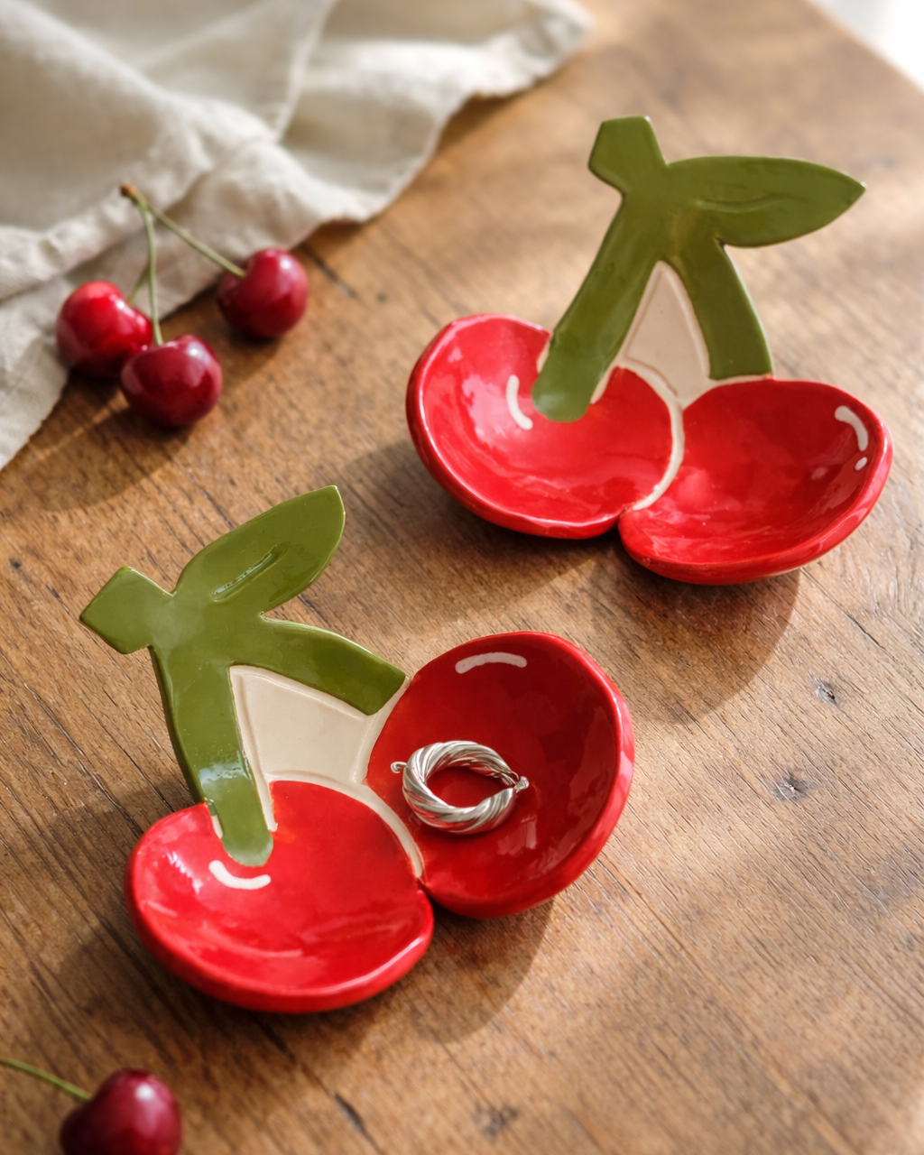Ceramic cherry trinket dish with silver ring on rustic wooden table with fresh cherries and linen cloth, fruit shaped jewelry holder for bedside or vanity decor