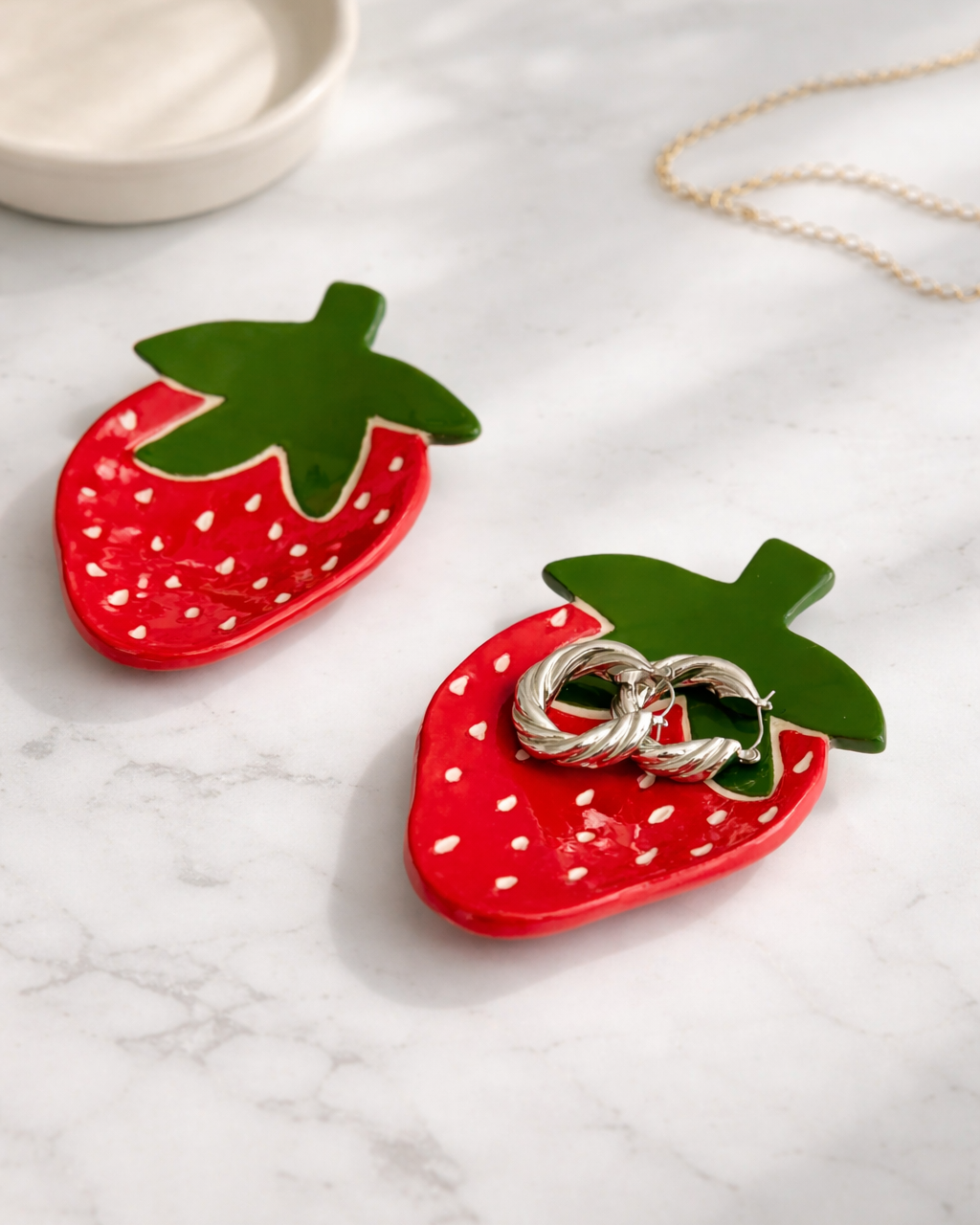 Ceramic strawberry jewelry dish holding hoop earrings on marble surface, handmade fruit trinket dish for vanity decor and jewelry organization