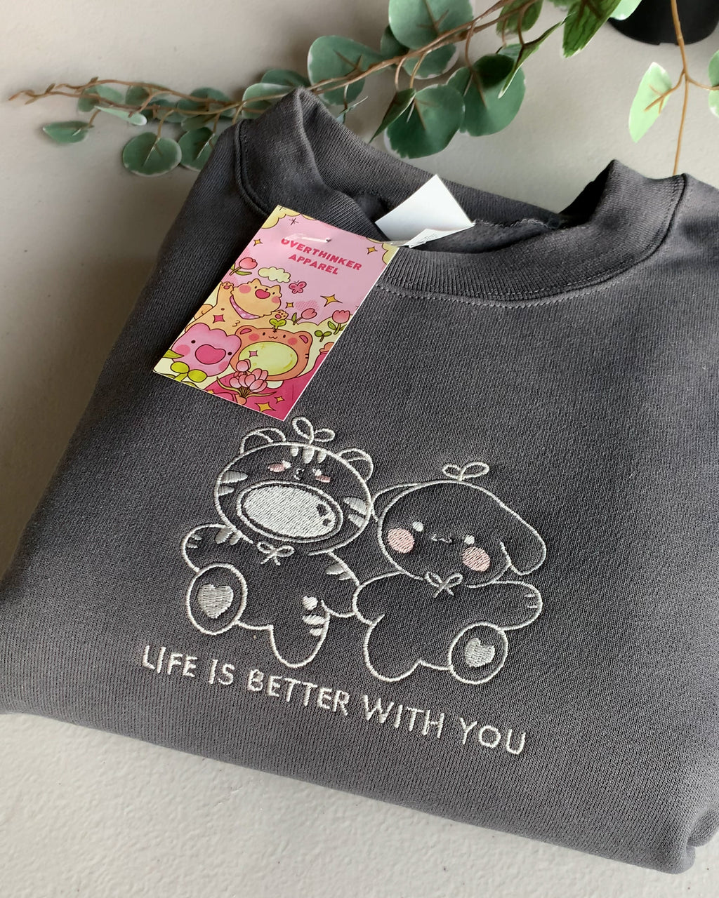 Close-up of a charcoal gray embroidered crewneck sweatshirt showing two cute characters and soft stitching detail.