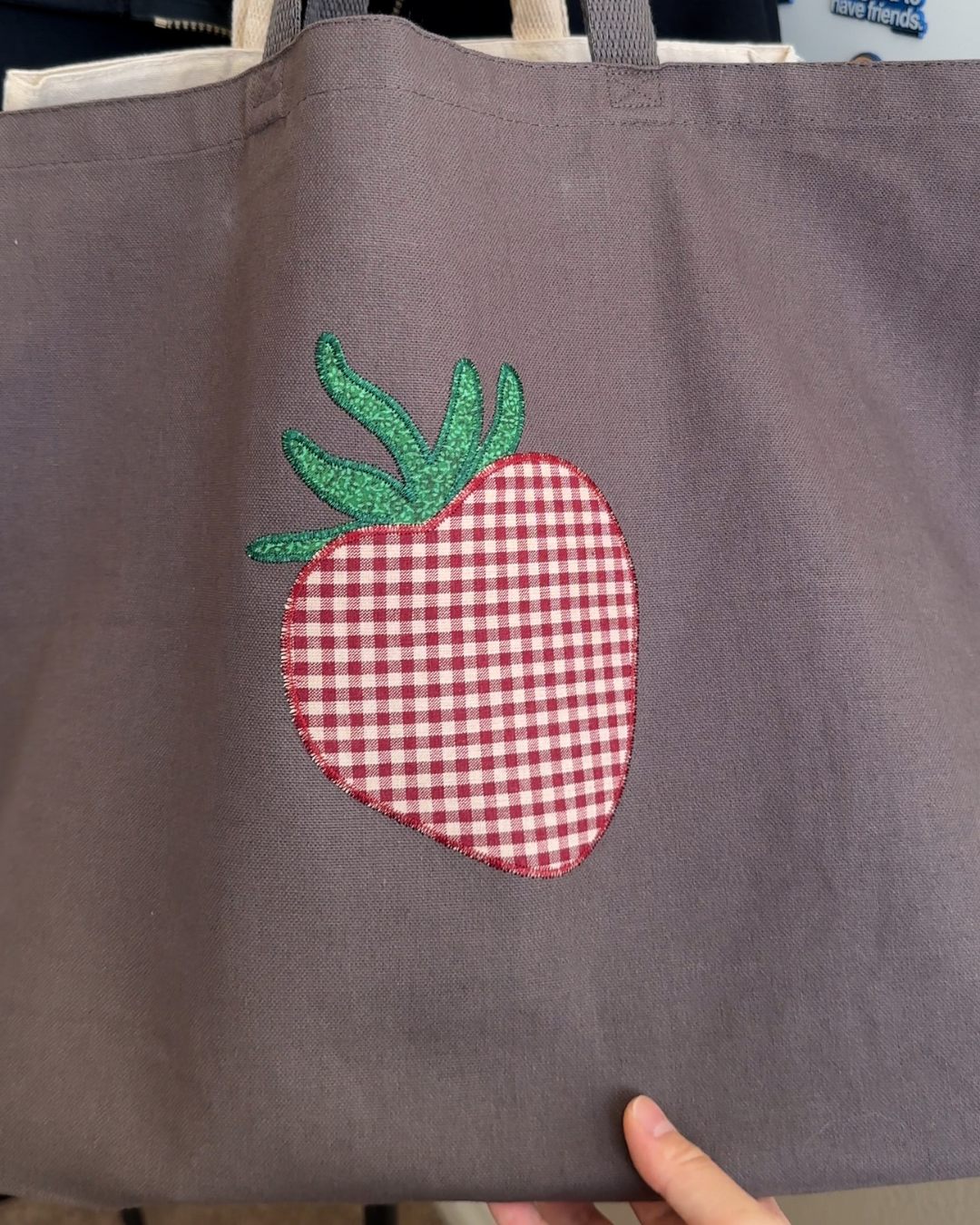Close up of strawberry embroidery on charcoal canvas tote with red gingham pattern and green stitched leaves