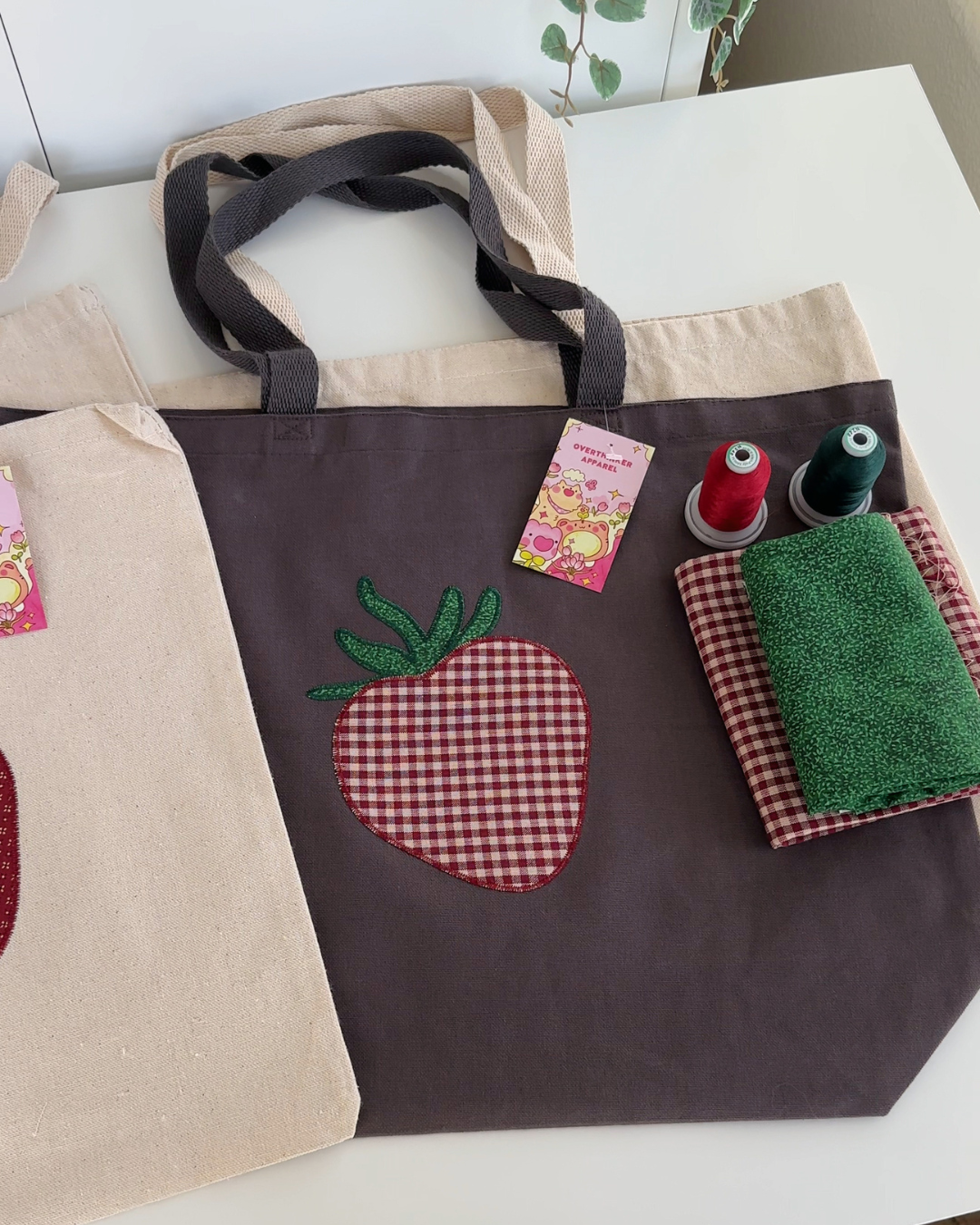 Charcoal canvas tote with strawberry embroidery next to thread spools and fabric, showing materials used in embroidered tote design