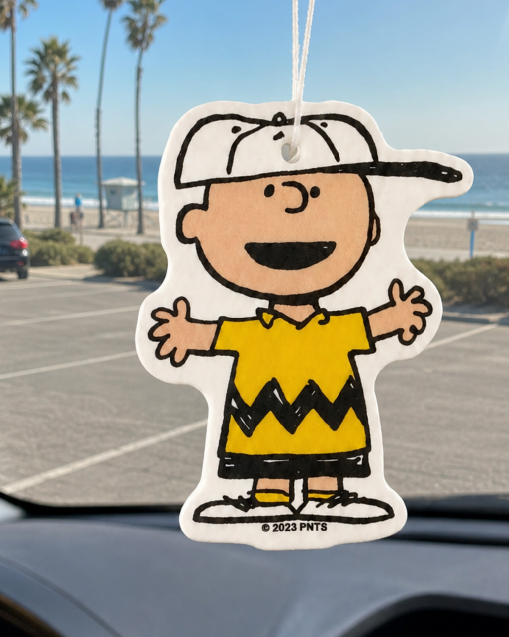 Charlie Brown air freshener with open arms and a big smile, hanging from a car mirror with a beach and palm trees in the background.