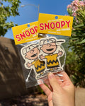 Hand holding two packaged Charlie Brown Morning Mist air fresheners against a sunny blue sky with greenery.