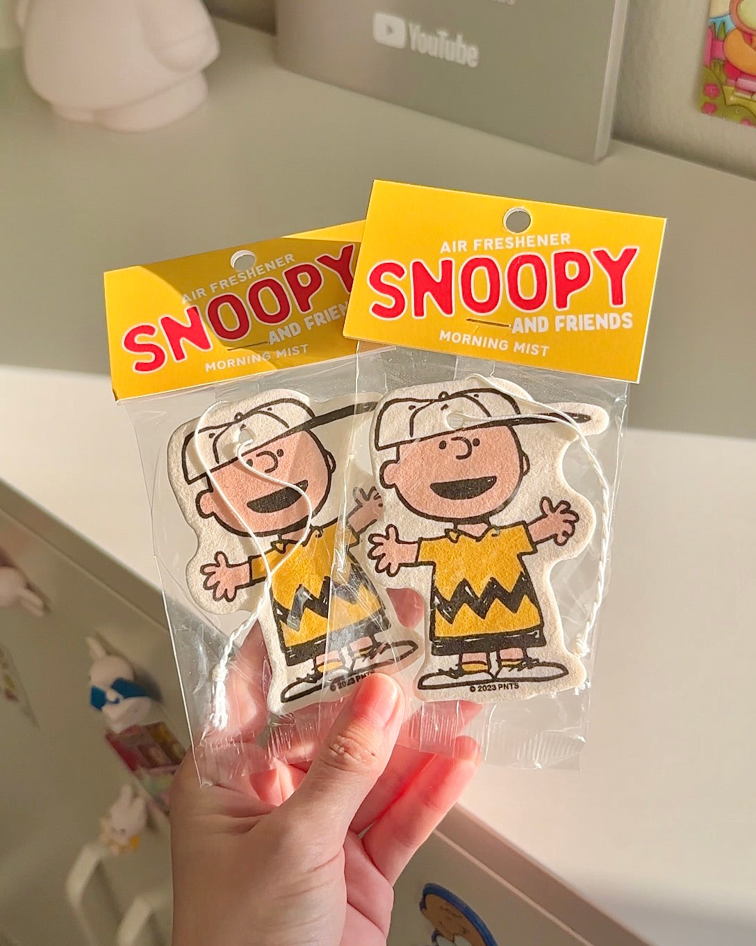 Two Charlie Brown air fresheners held in hand. Morning Mist scent with cheerful design and eco-friendly packaging. Peanuts-themed car decor.