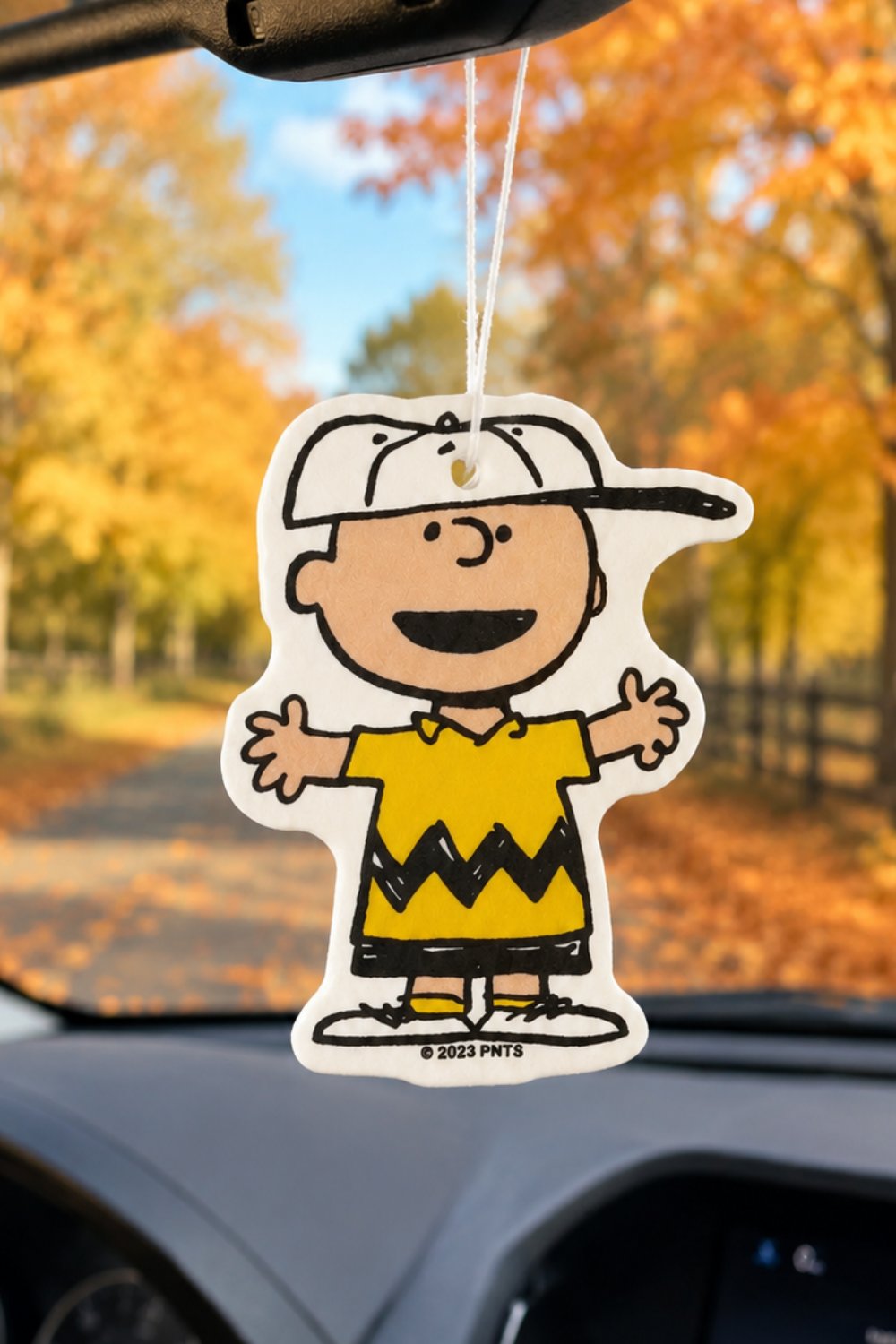 Charlie Brown car air freshener with arms wide open and a big smile, hanging from a rearview mirror with autumn trees outside.
